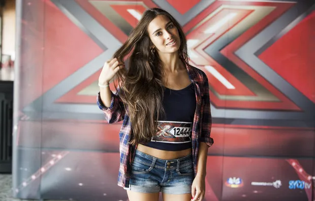 Clara Neto de camisa aos quadrados e calções de ganga, a mexer no cabelo, junto ao cenário do Factor X