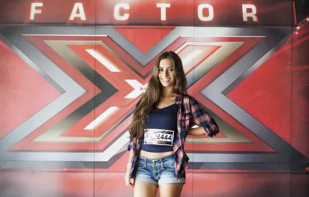 Clara Neto sorridente, com o dístico de concorrente ao peito, em frente ao cenário do Factor X