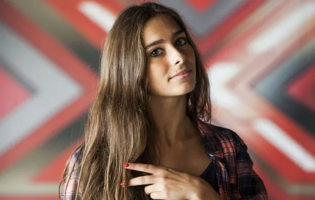 Retrato de Clara Neto, de cabelo castanho comprido, em frente ao X vermelho do cenário do Factor X