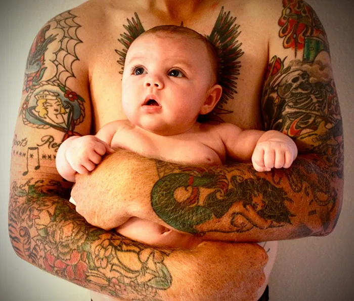 Bebé de olhos muito abertos seguro pelos braços tatuados do pai, com uma tatuagem de asas no peito