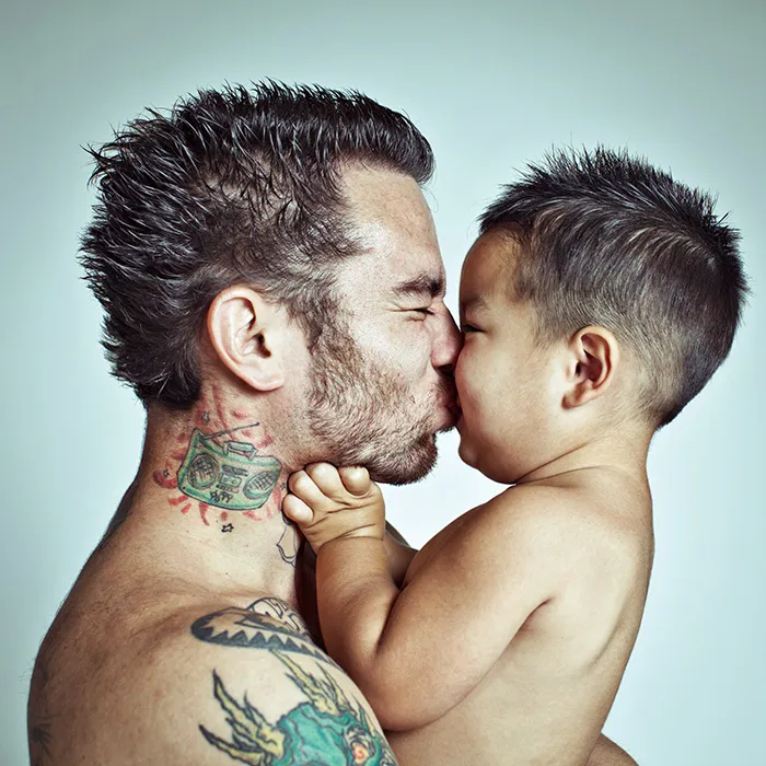 Pai com tatuagens no pescoço dá um beijo ao filho pequeno, cara com cara