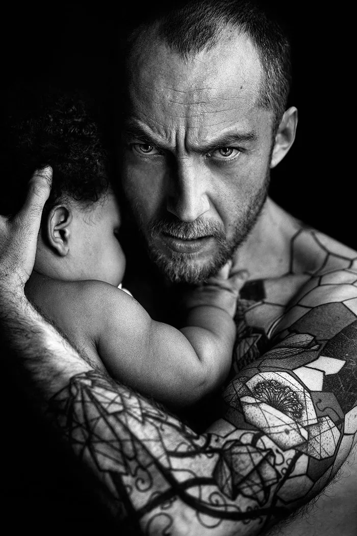 Pai de expressão séria segura o bebé contra o peito, com o braço tatuado, a preto e branco