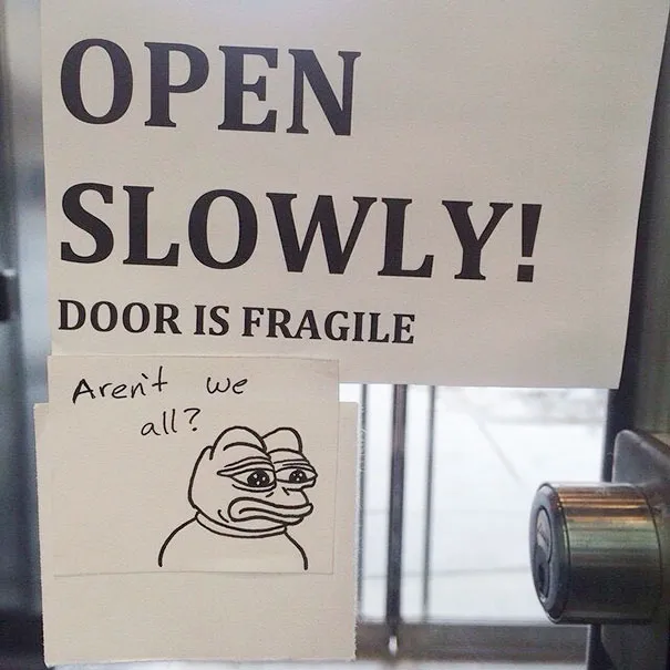 Aviso numa porta a dizer abre devagar, a porta é frágil, com um papel por baixo a perguntar se não somos todos e o desenho de um sapo triste