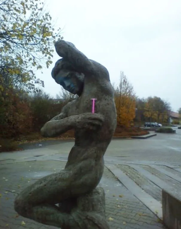 Estátua de uma figura nua a levantar o braço, com uma lâmina de barbear cor-de-rosa na mão