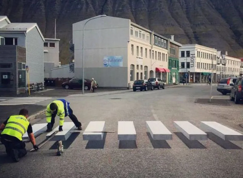 Dois trabalhadores de colete refletor a pintar a passadeira 3D numa rua de Ísafjörður, com casas e uma montanha ao fundo