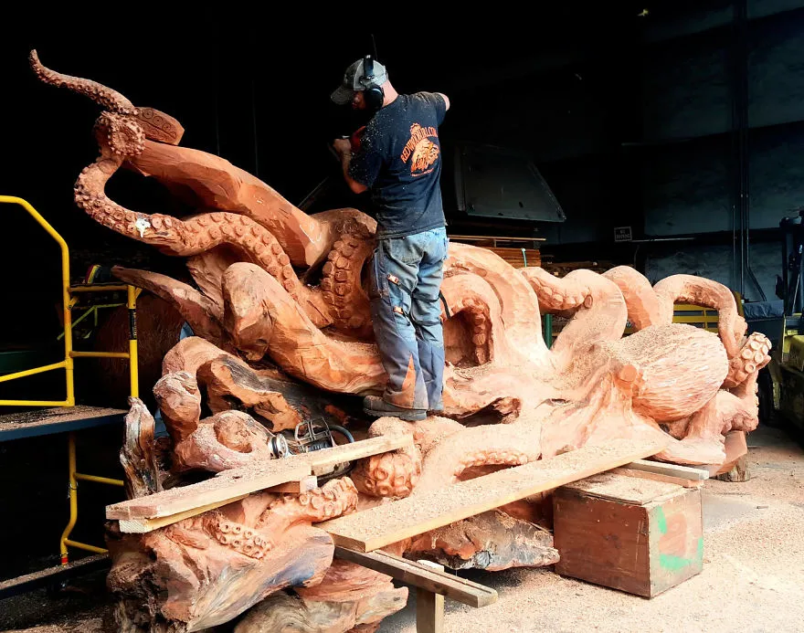 O escultor de pé em cima da escultura, a trabalhar a ponta de um tentáculo com proteção nos ouvidos