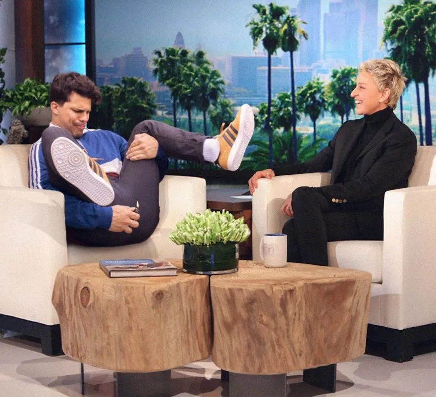 Average Rob de pernas encolhidas numa poltrona de um programa de entrevistas, diante da apresentadora