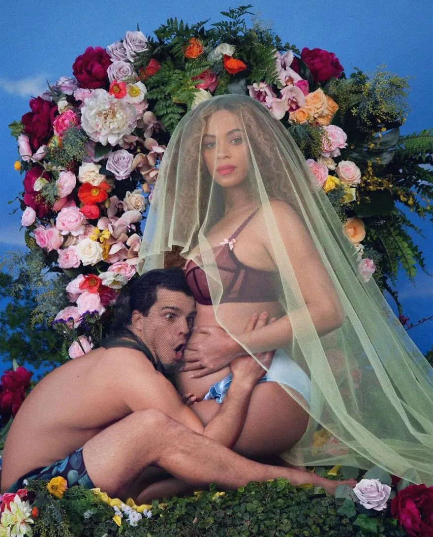 Average Rob encostado à barriga de uma mulher grávida de véu, diante de uma parede de flores