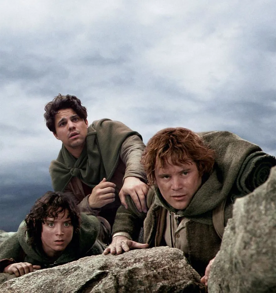 Average Rob espreita assustado por trás de uma rocha, ao lado de dois hobbits numa cena de fantasia