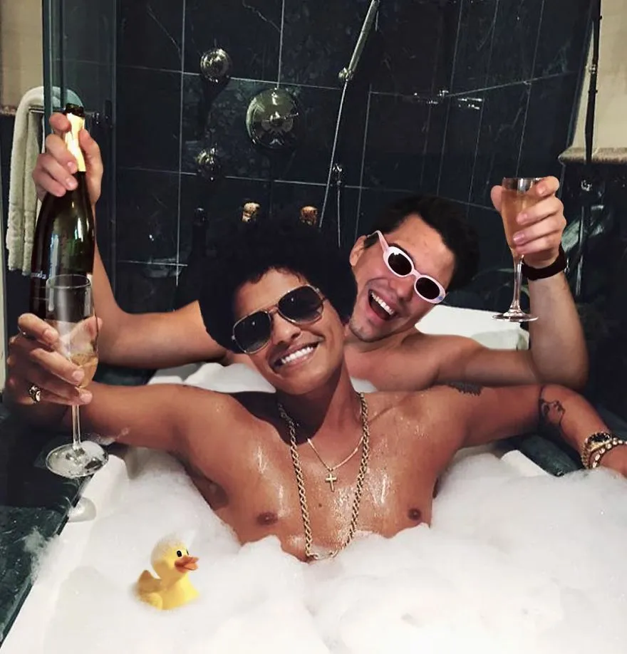 Average Rob numa banheira de espuma com um copo de champanhe, ao lado de um cantor de óculos escuros