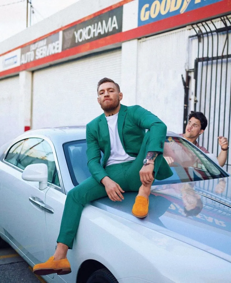 Homem de fato verde sentado no capô de um carro branco, com Average Rob a acenar ao fundo