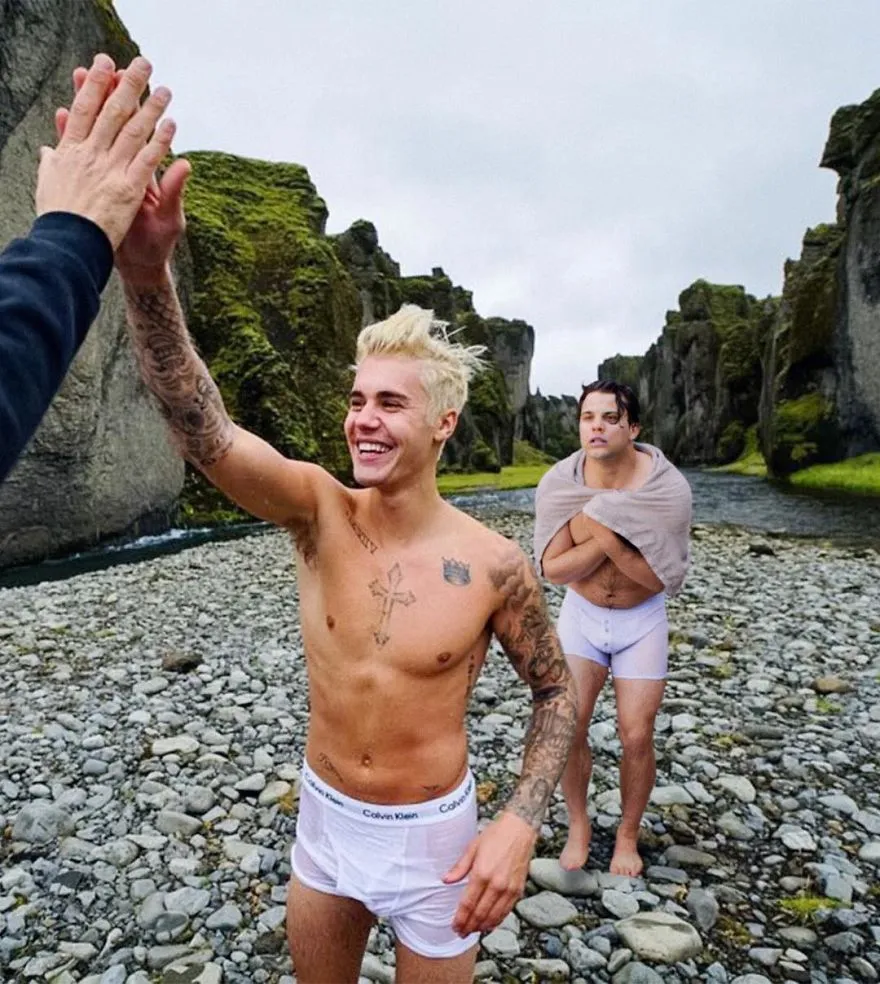 Jovem tatuado em cuecas brancas dá um mais cinco à beira de um rio, com Average Rob a tremer de frio ao fundo