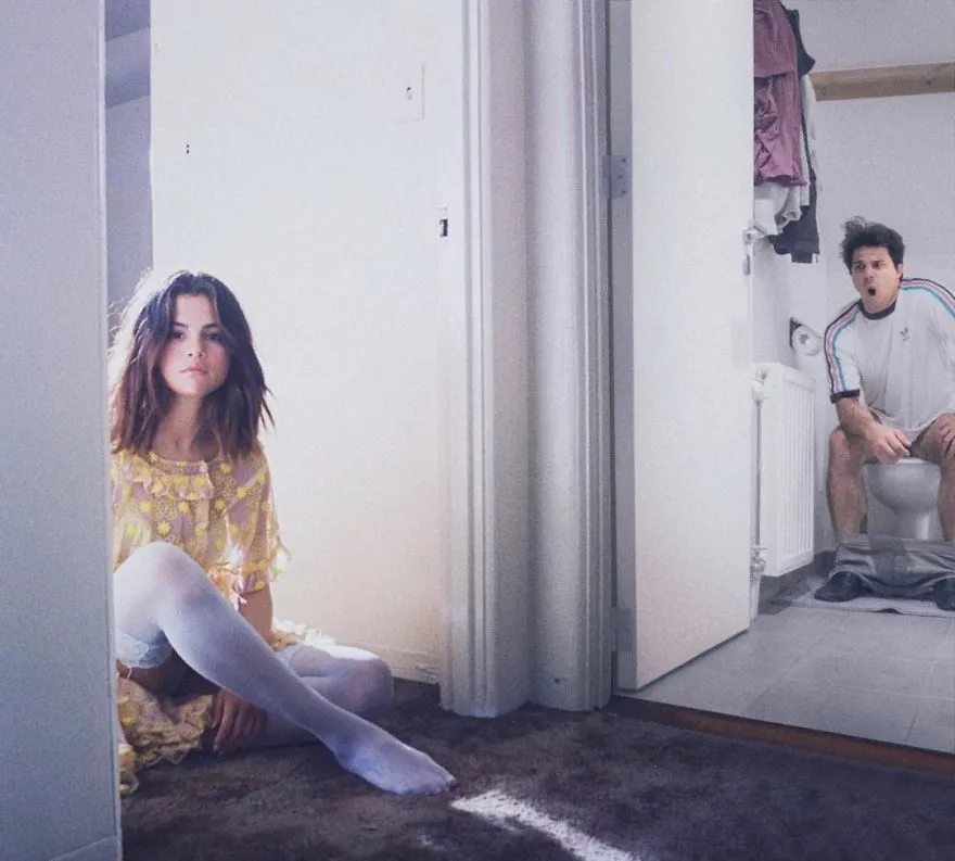 Mulher sentada no chão de um quarto enquanto, pela porta aberta, se vê Average Rob sentado na sanita