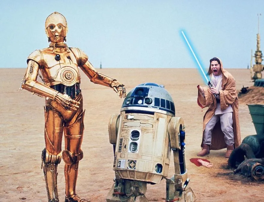 Os robôs C-3PO e R2-D2 no deserto, com Average Rob atrás deles de túnica e sabre de luz