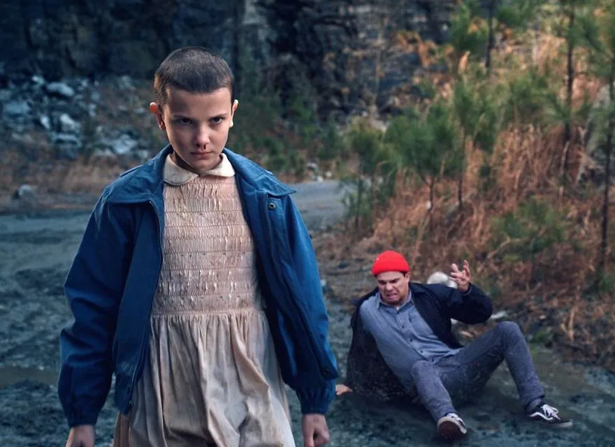 Rapariga de cabelo rapado e vestido cor-de-rosa, numa cena inspirada na série Stranger Things, com Average Rob a cair para trás
