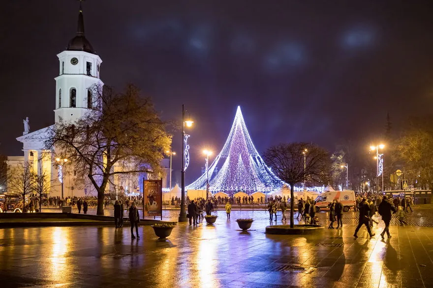 Árvore de Natal de Vilnius vista da praça molhada, com o campanário e o reflexo das luzes no chão
